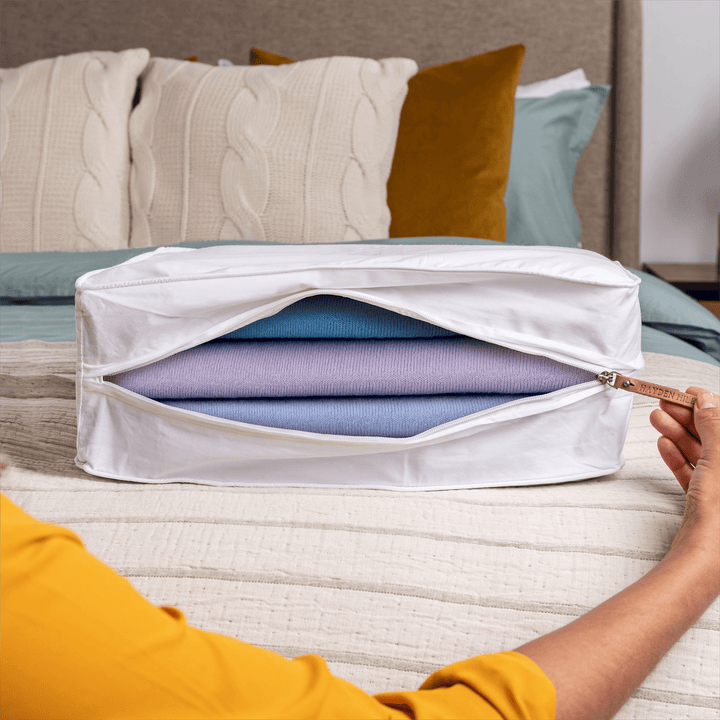 a close up of a large storage bag sitting on a bed begin unzipped with coloured sweaters stored inside