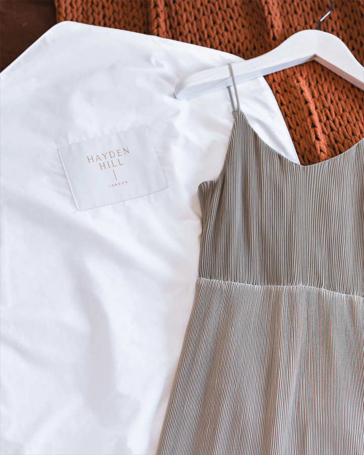 a hanging garment bag lying on a woollen blanket next to a grey summer dress with shoestring straps