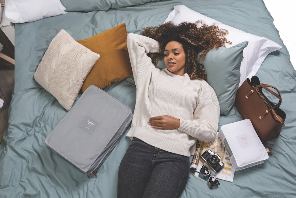 a woman lying on her bed surrounded by cushions, a purse and a two storage bags, one in grey and one in white
