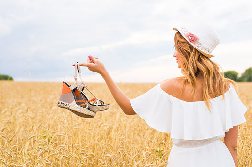 a woman standing in a wheat field wearing a white dress and holding a pair of sandals