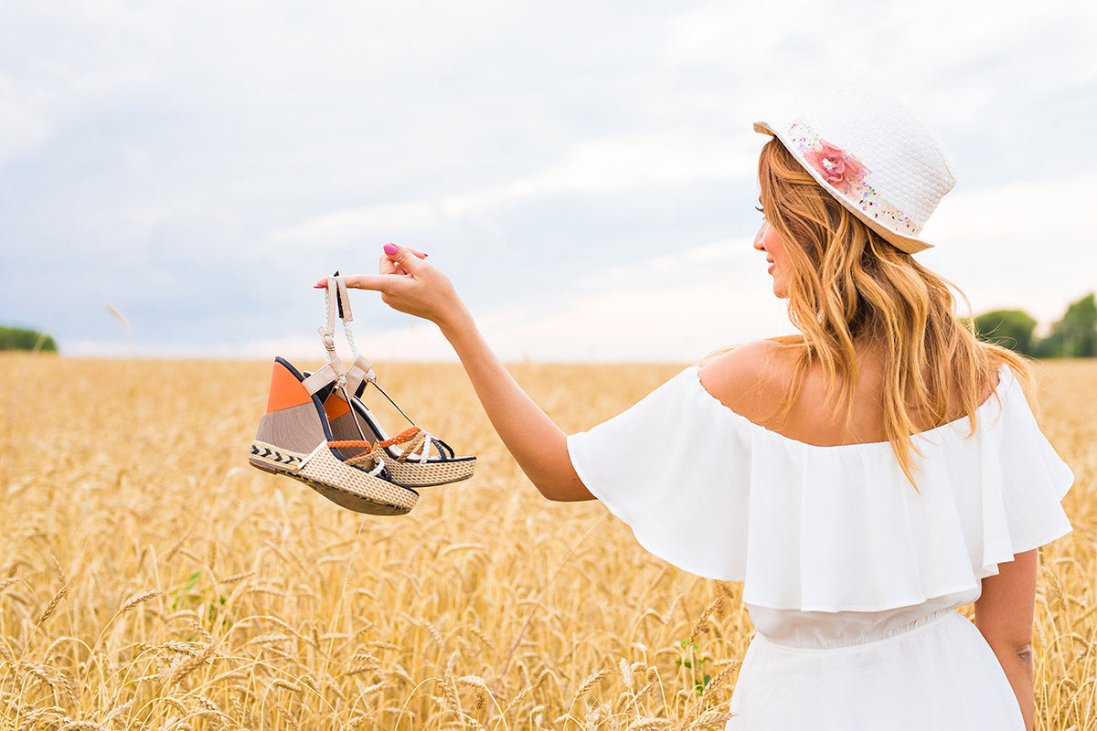 a woman standing in a wheat field wearing a white dress and holding a pair of sandals