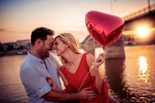 a couple embracing by the river at sunset, the woman is holding a red heart shaped balloon