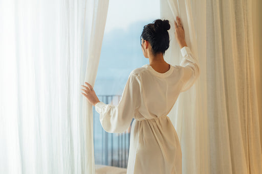 a woman in a soft cream dress holding open the curtains and looking out at the view