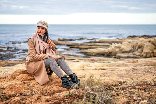 a beautiful woman wrapped warmly in a camel coat, large scarf and leggings sitting by the sea