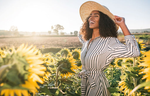 a laughing woman standing in a field of sunflowers wearing a stripy dress and sunhat