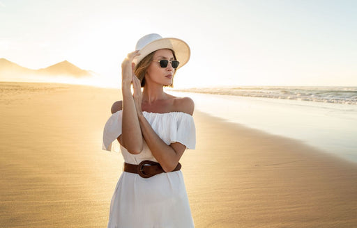 a woman on the beach wearing a white off the shoulder dress, brown belt, hat and sunglasses