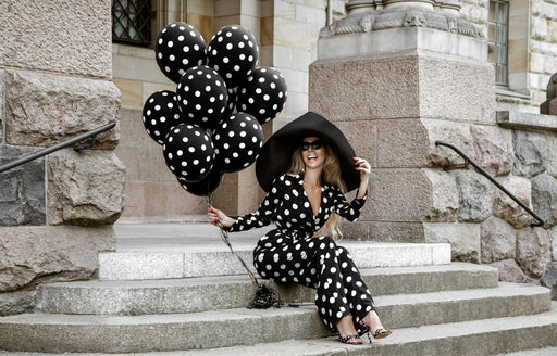 a woman wearing a black and white polka dot jumpsuit and large brim hat, holding black and white polka dot balloons