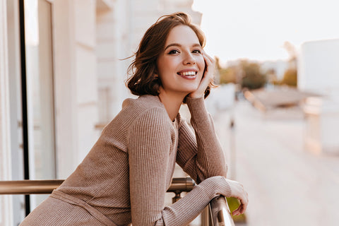 a woman wearing a light brown ribbed wool sweater leaning on her balcony railing