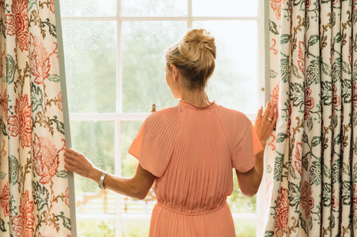 a woman wearing a peach coloured silk dress looking out of the window through parted curtains