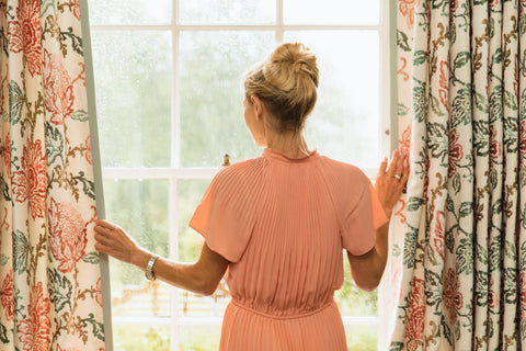 a woman wearing a peach coloured silk dress looking out of the window through parted curtains