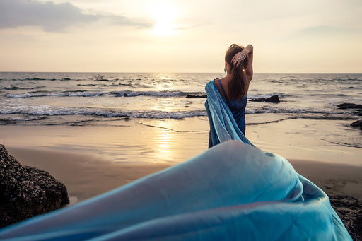 a woman with a beautiful swathe of blue silk standing by the ocean at sunset