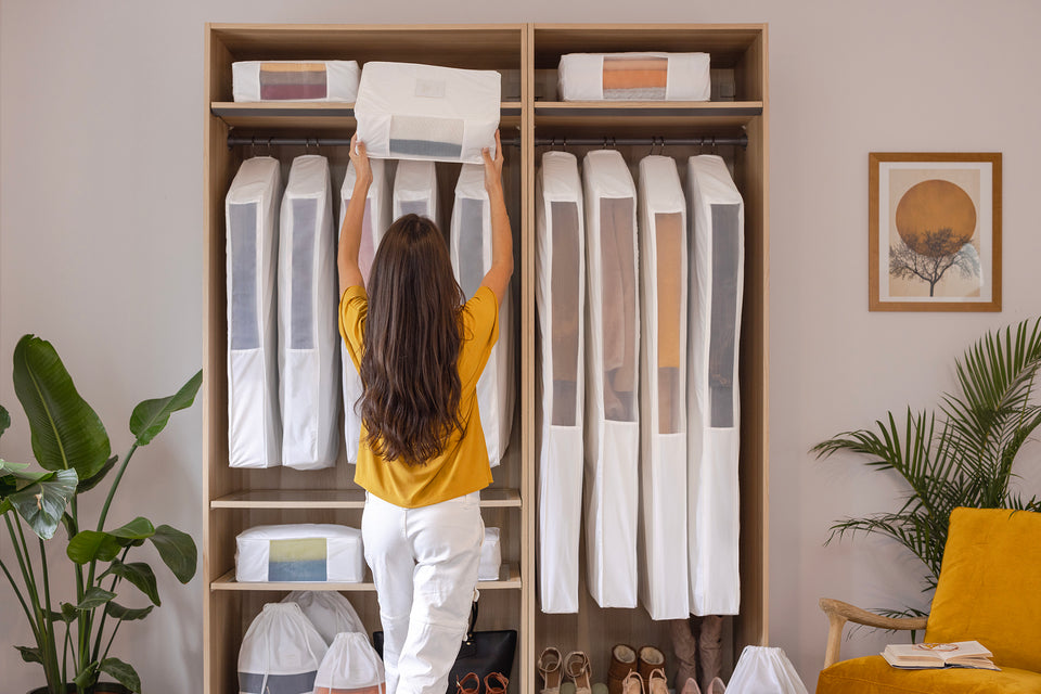 A woman putting an extra large Hayden Hill storage cube into an organised closet which is filled with hanging bags for clothes, short hanging garment bags, good-sized packing cubes, and multiple dust bags for purses, handbags or shoes.