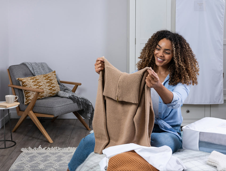 A woman holding up her camel colored wool sweater, having opened her Hayden Hill sweater storage bag