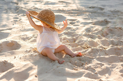 a baby in a white dress and a straw hat sitting on the beach in the sunshine