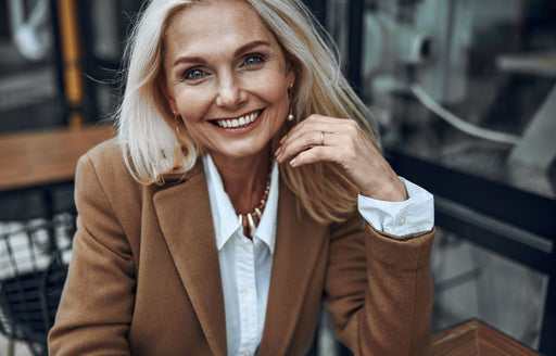a beautiful blonde older woman wearing a classic white shirt and a camel coloured overcoat