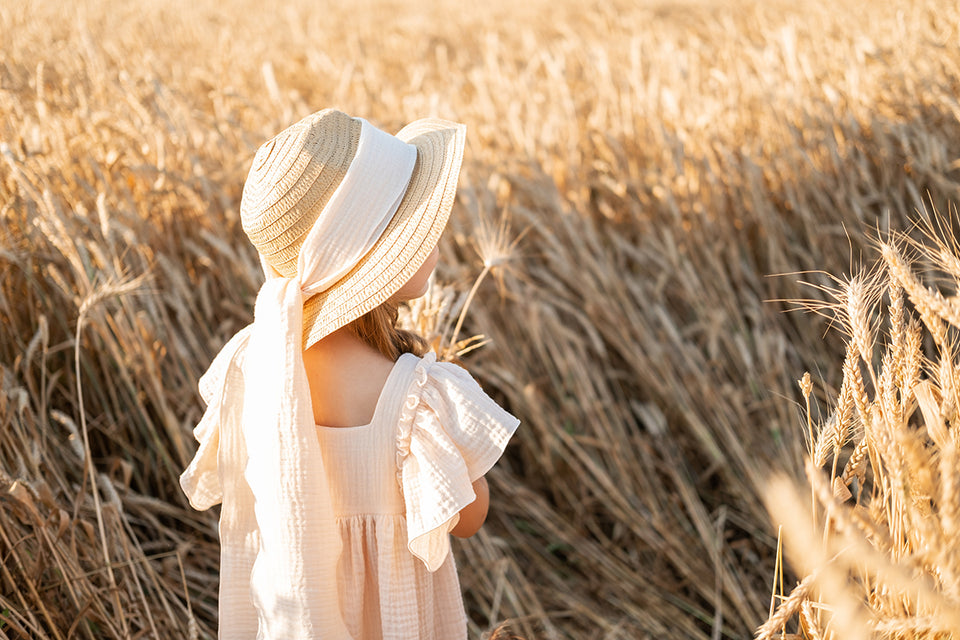 a little girl in a sunny wheat field wearing an unbleached muslin dress and a straw hat