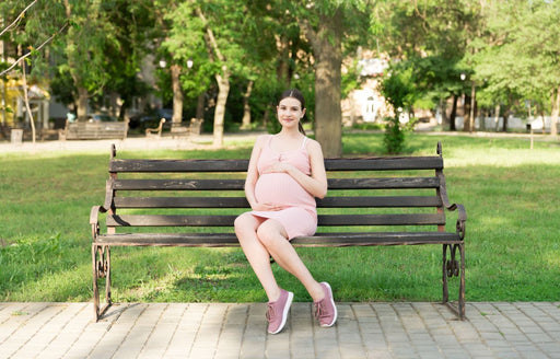 a pregnant woman in a pale pink dress and sneakers sitting on a park bench