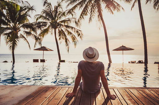 a relaxed man sitting on the edge of a pool looking out at the sea and palm trees