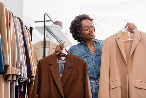 a woman holding up a dark brown blazer and a caramel colored blazer on white wooden hangers