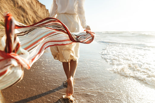  a woman walking on the beach wearing floaty clothing and a striped scarf flowing behind her