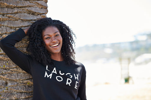 a woman wearing a ‘Laugh More’ black sweatshirt looking relaxed leaning on a palm tree