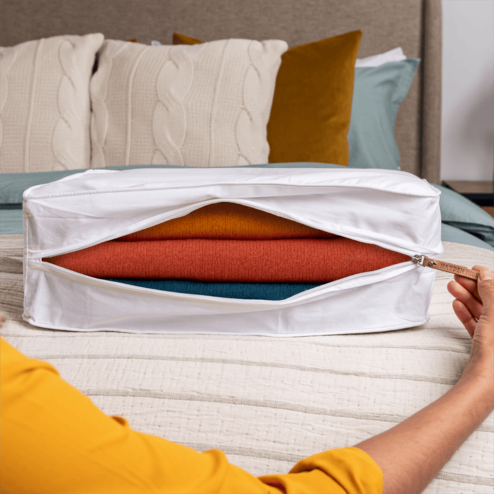 a close up of an extra large storage bag sitting on a bed begin unzipped with coloured sweaters stored inside