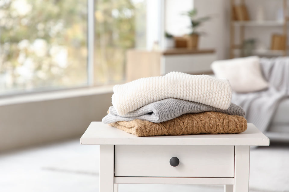 an simple assortment of sweaters piled onto a white bedside table